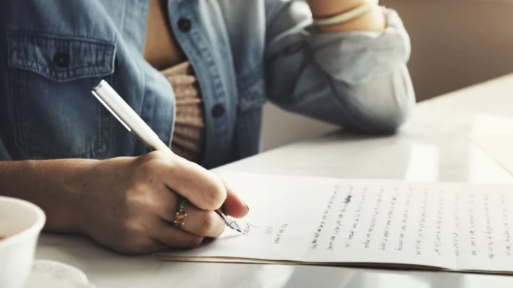 a woman sitting at a table writing on a piece of paper.
