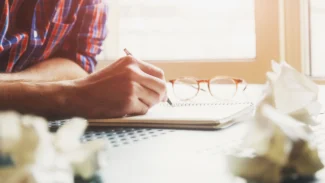 a person writing on a notebook with a pair of glasses on top of it